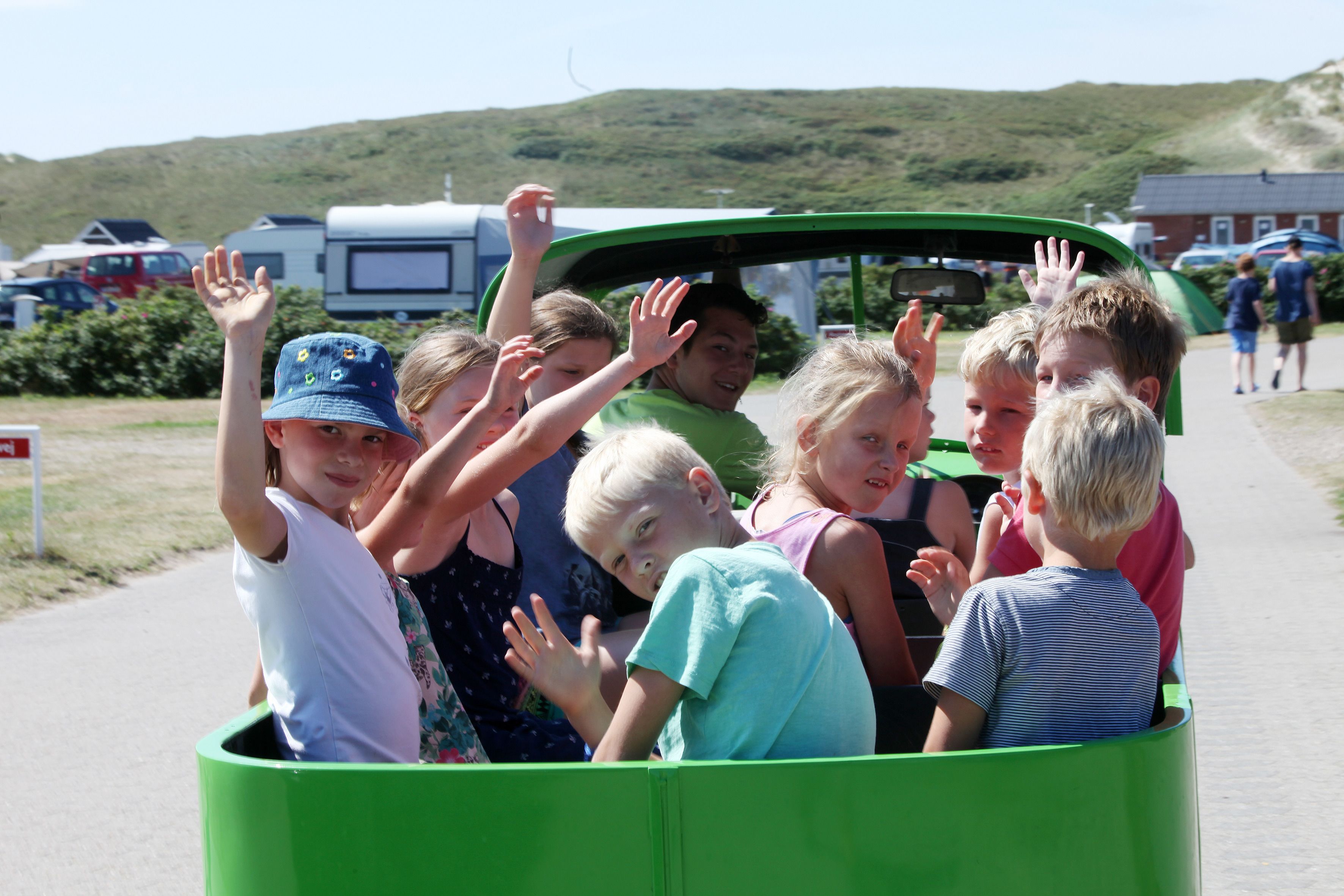 A group of children in a green vehicle with its top open, waving their hands.