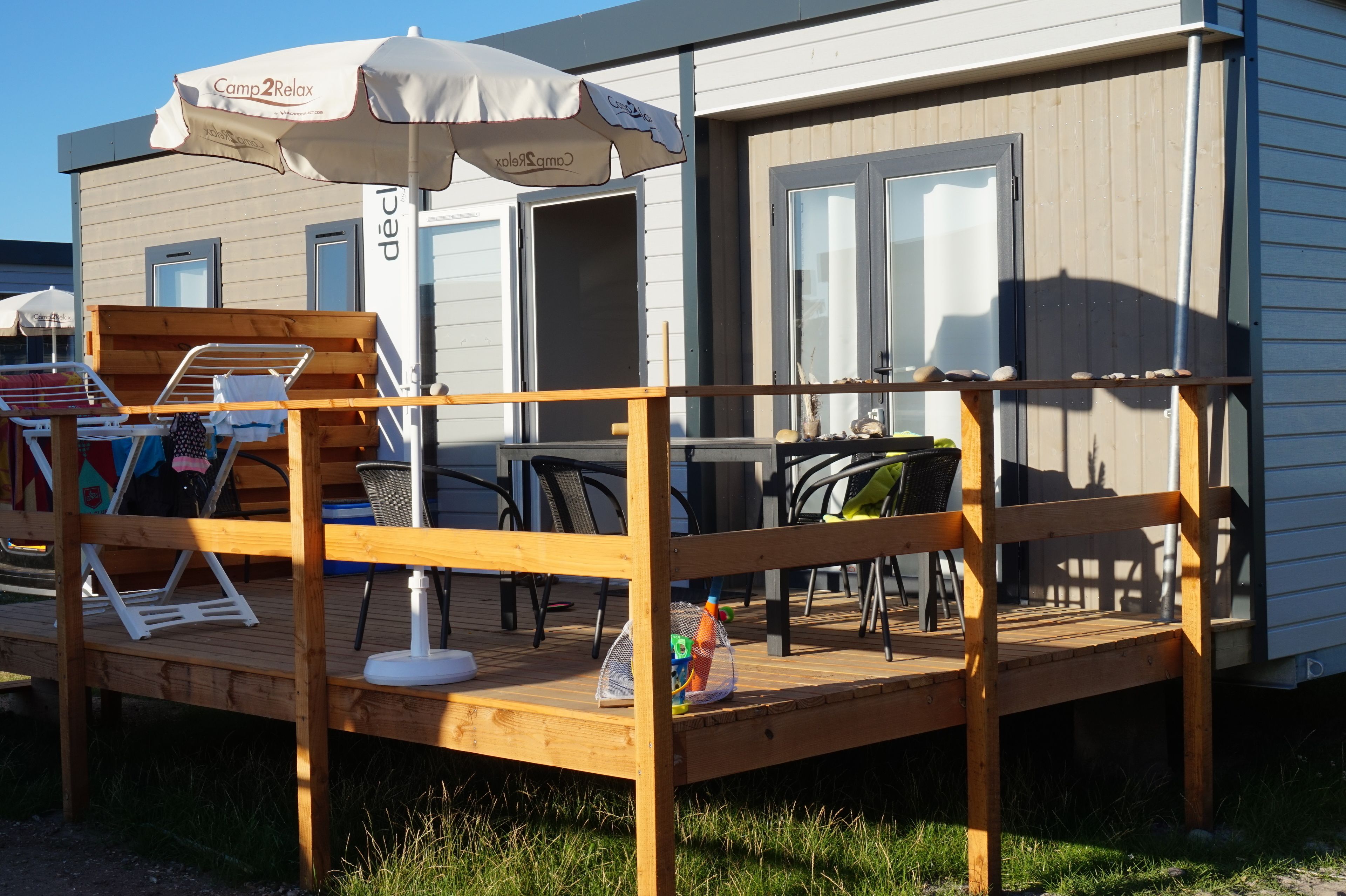 Una terraza de madera con mobiliario exterior, un espacio de estar techado y un edificio con una puerta y ventanas.
