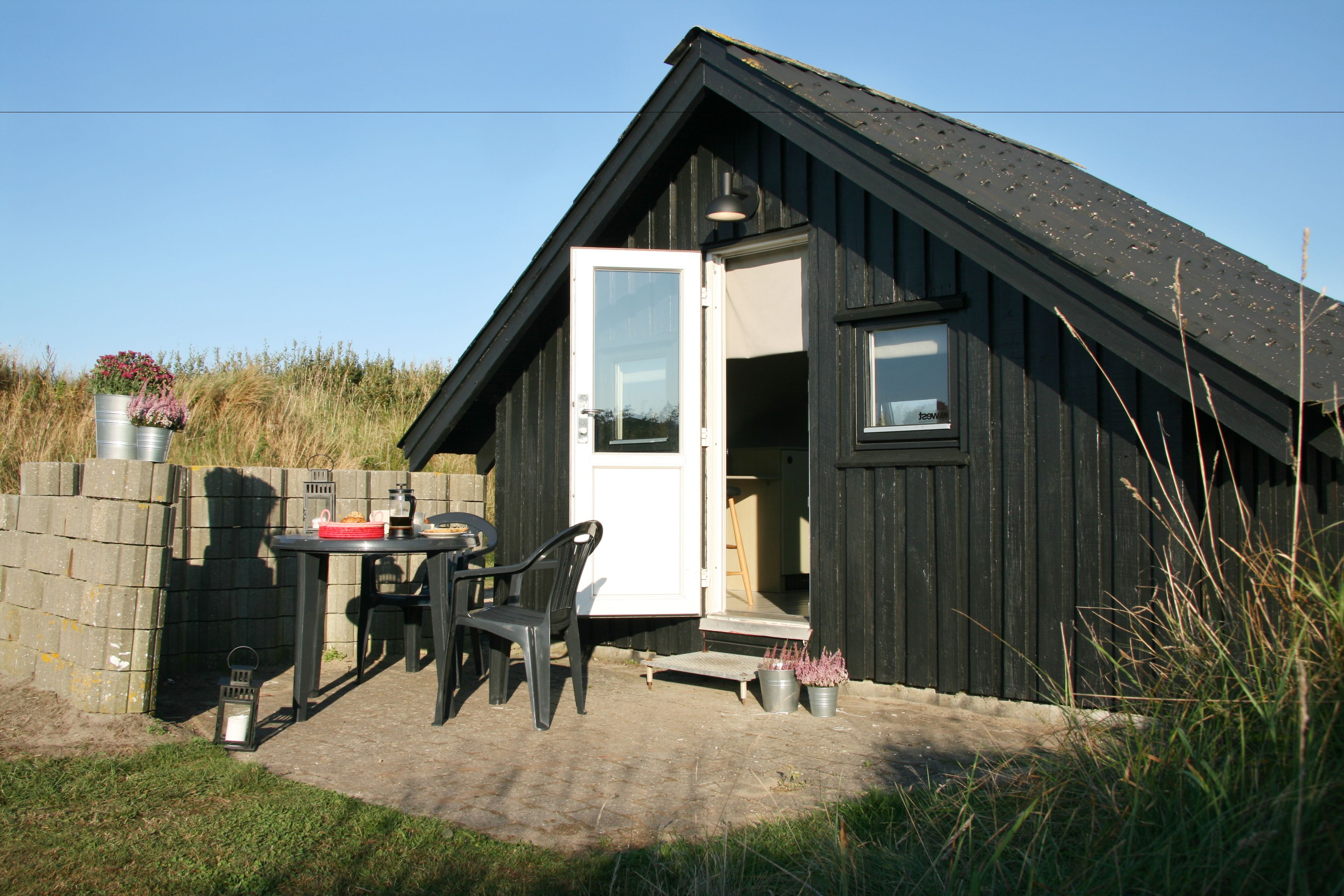 Eine kleine Holzhütte mit offener Tür, einem Tisch mit Stühlen und einem Blumentopf davor.