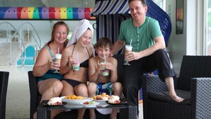 Eine vierköpfige Familie sitzt an einem Tisch im Freien, mit Getränken und Speisen, im Hintergrund ist ein Pool und eine Liegefläche zu sehen.
