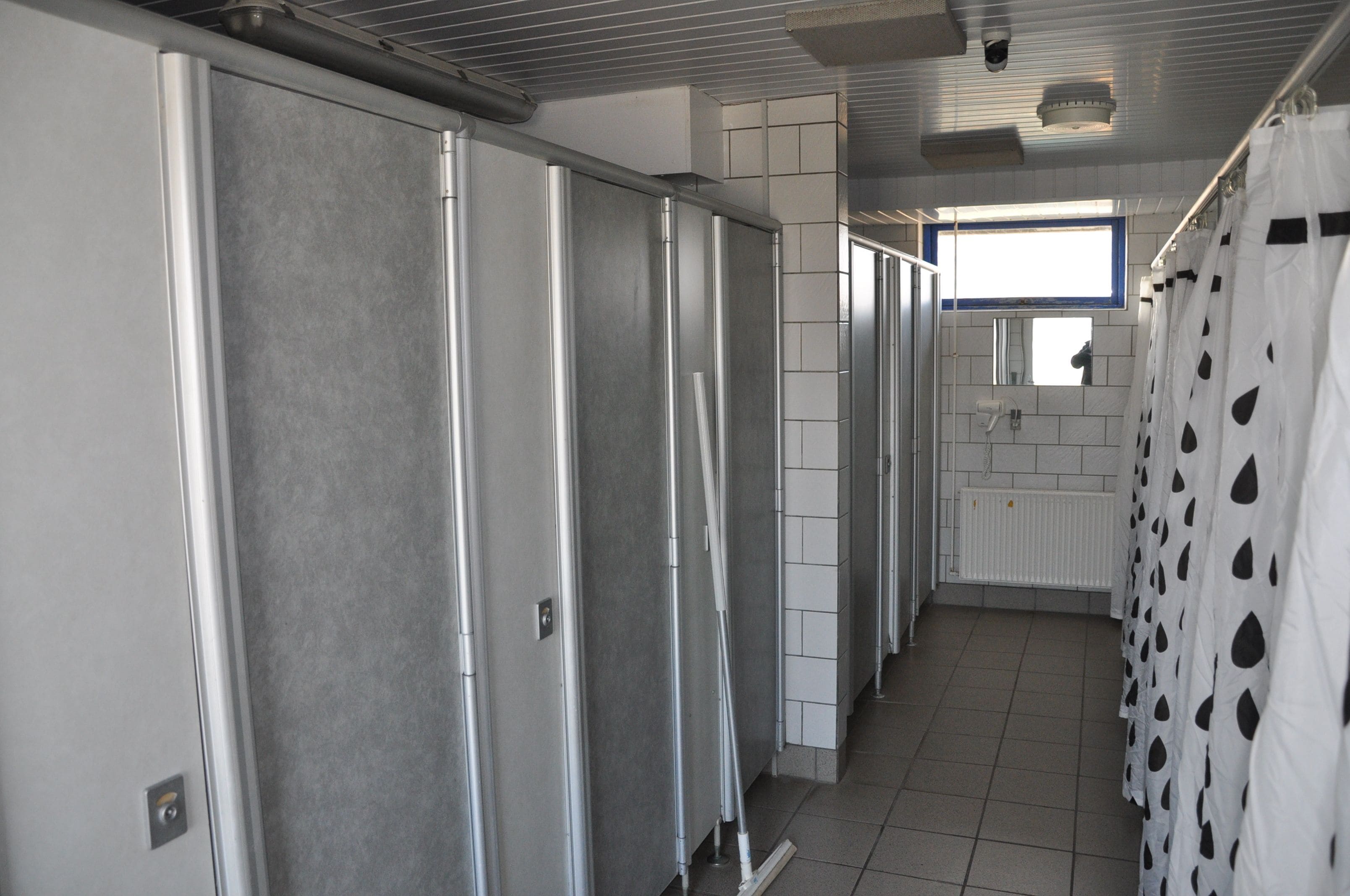 A corridor with metal lockers and a tiled floor, leading to a bathroom with a window and a shower curtain with a pattern.