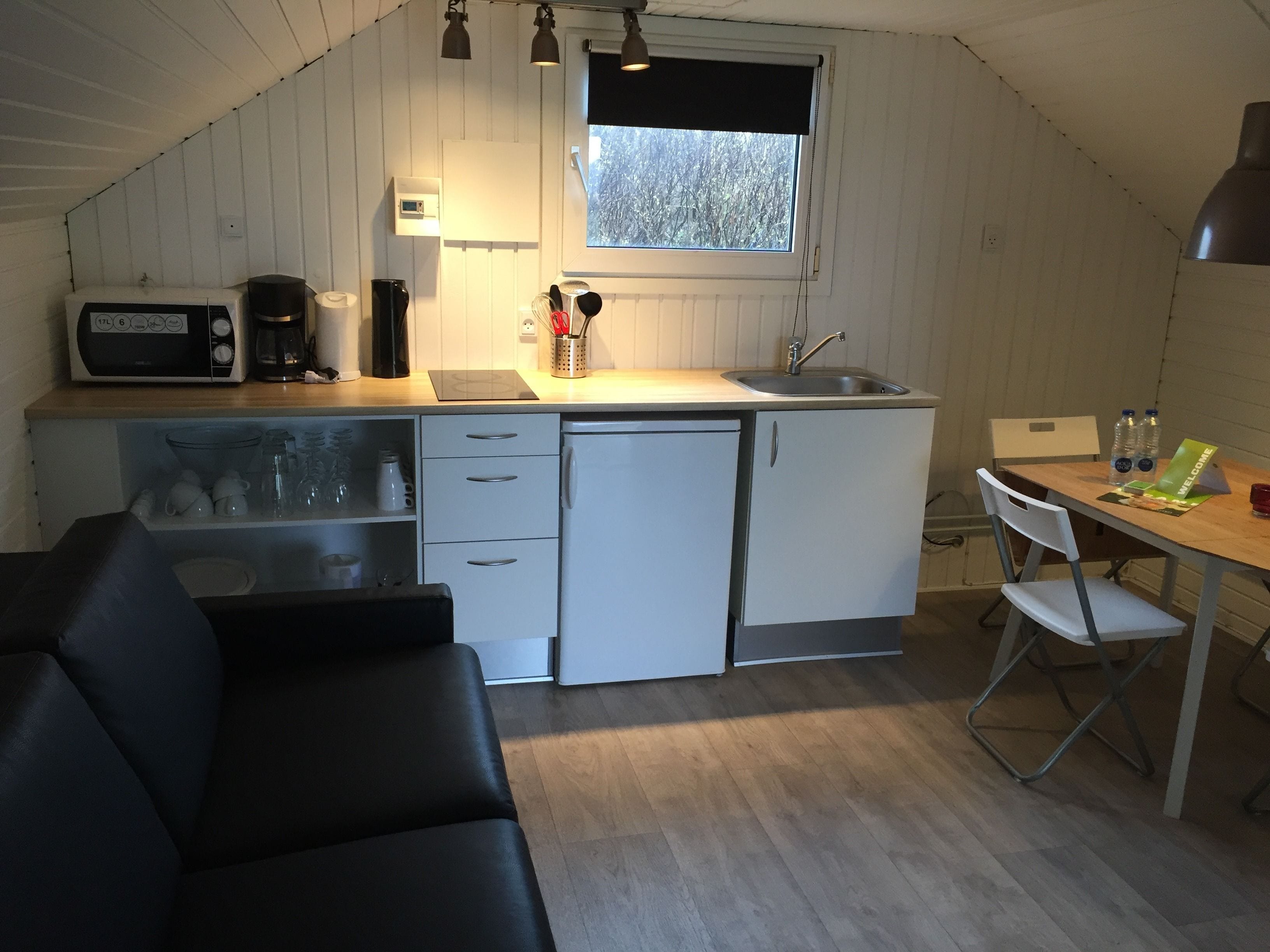 A compact kitchen with a microwave, coffee maker, and sink. There is a small refrigerator, a dining table with chairs, and a black leather couch.