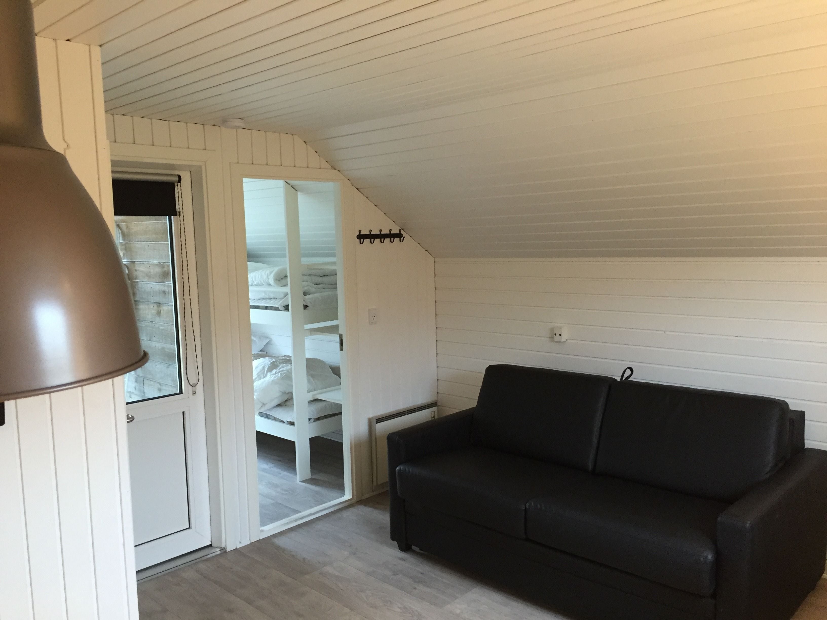 Een kamer met een zwarte leren bank, een witte deur die leidt naar een kast met hangende kleding, en een schuine plafond.