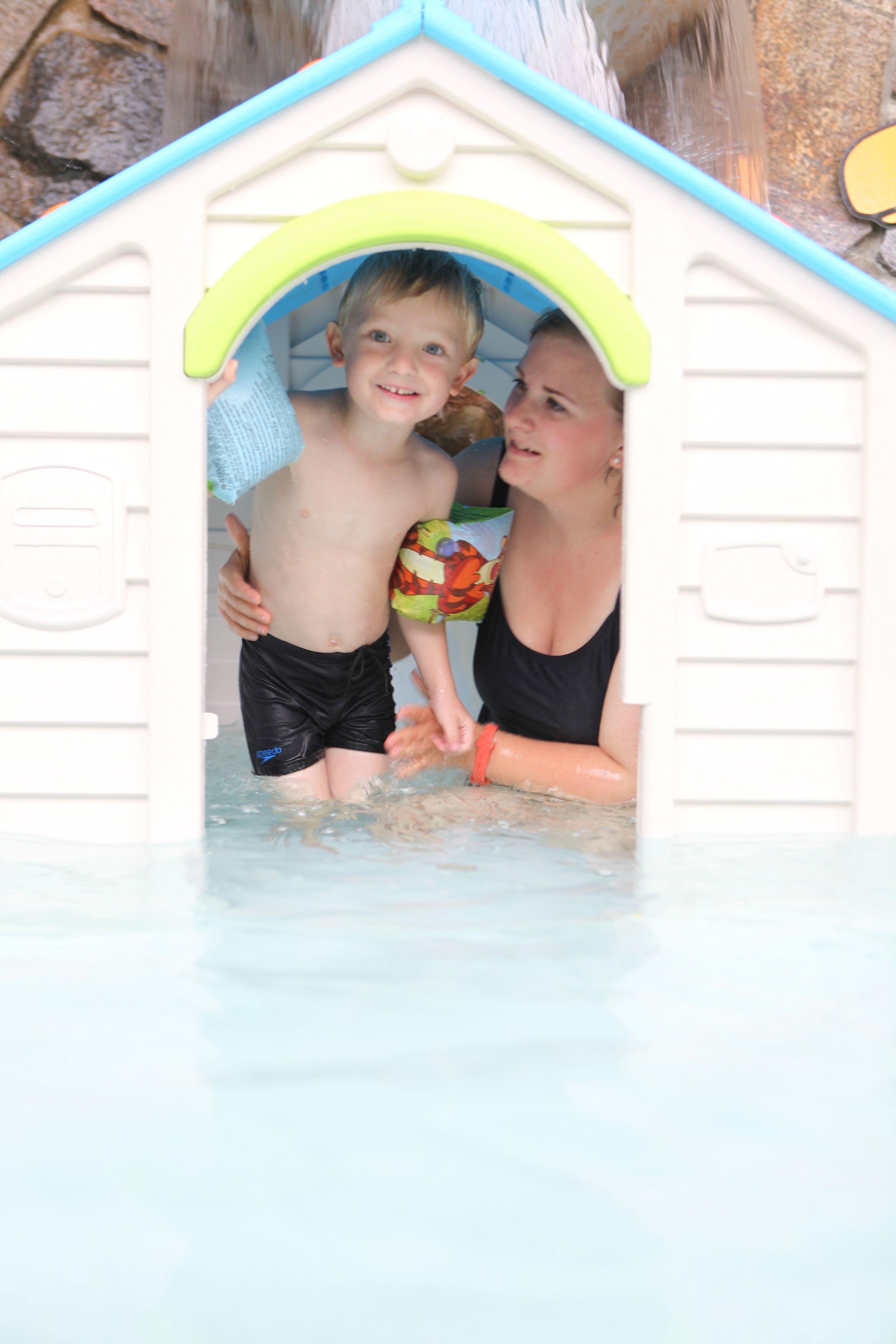 A child and an adult in a small, white, house-shaped pool with a blue roof.