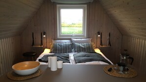 Ein gemütliches Schlafzimmer im Dachgeschoss mit holzgetäfelter Schräge, einem großen Fenster, einem Bett mit gestreiften Kissen, einem Nachttisch mit Schale und Kerzen sowie einer Kaffeemaschine.