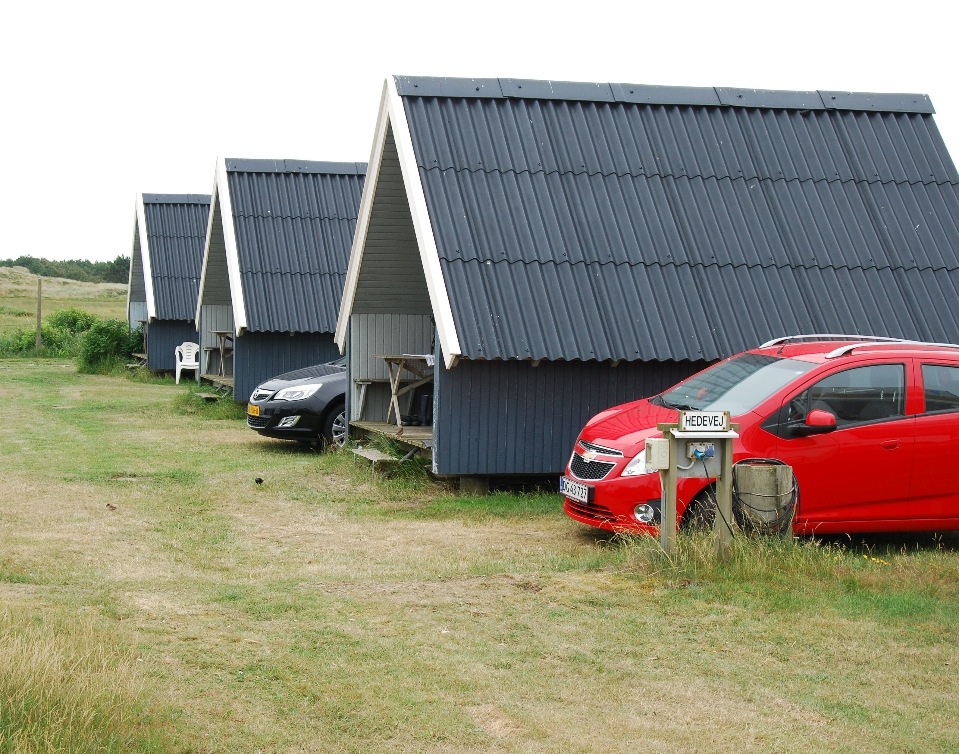 En række små, mørke hytter med skrå tage, hvor der er parkeret en bil foran hver.