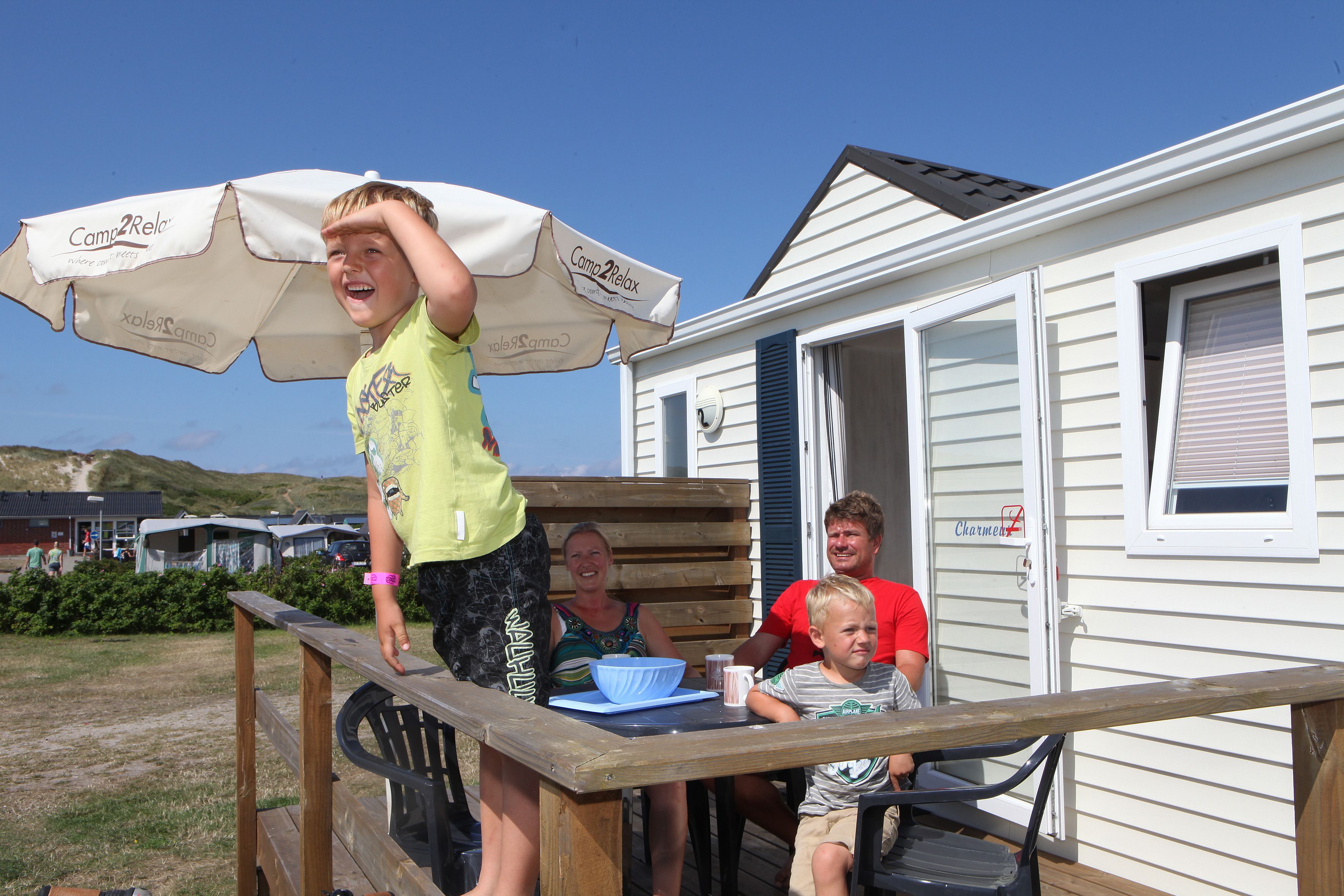 Une famille est assise à une table à l’extérieur, sous un parasol blanc, sur une terrasse en bois, avec une maison mobile en arrière-plan.