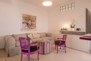 A living room with a beige sofa, two pink chairs, a striped ottoman, a painting on the wall, a small table with candles and a plant, and a TV on a stand. - Iron Bridge Accomodation (Rome)
