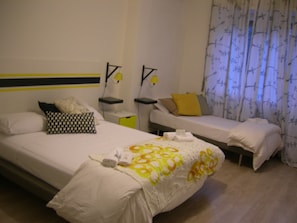 Una habitación de hotel con una cama grande, dos camas más pequeñas y una mesita de noche con una lámpara.