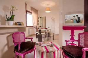 A room with pink chairs, a striped ottoman, a television, a potted plant, and a bookshelf. - Iron Bridge Accomodation (Rome)