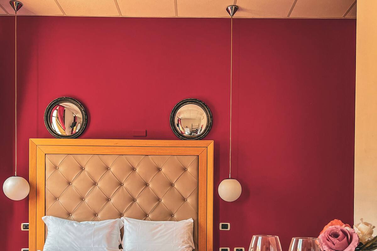Una cama bien hecha con cabecera de madera, dos mesitas de noche y una pequeña mesa con dos vasos y un jarrón de flores.