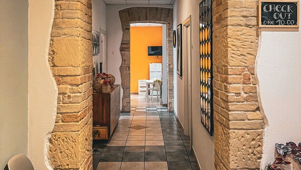 A hallway with exposed brick walls, a check-out sign, and a tiled floor leading to a room with a bright orange wall. - Chambres du Monde (Cagliari)