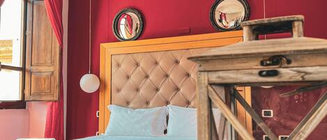 A bed with a wooden frame and a red bedspread, a wooden nightstand with a glass container, and a red wall with two round mirrors.