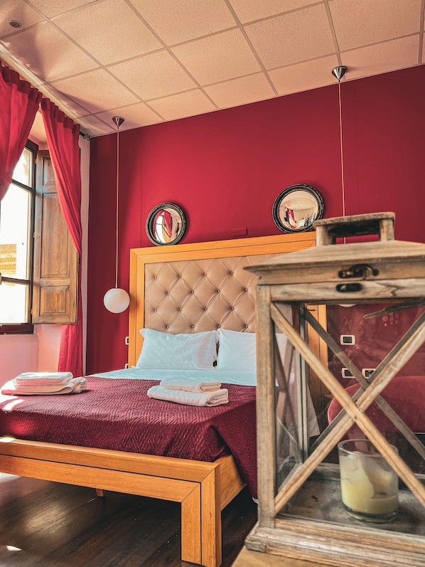 A bed with a wooden frame and a red bedspread, a wooden nightstand with a glass container, and a red wall with two round mirrors.