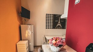 A compact hotel room with a wooden floor, a dining table with chairs, a refrigerator, a television mounted on the wall, and a red accent wall. - Chambres du Monde (Cagliari)