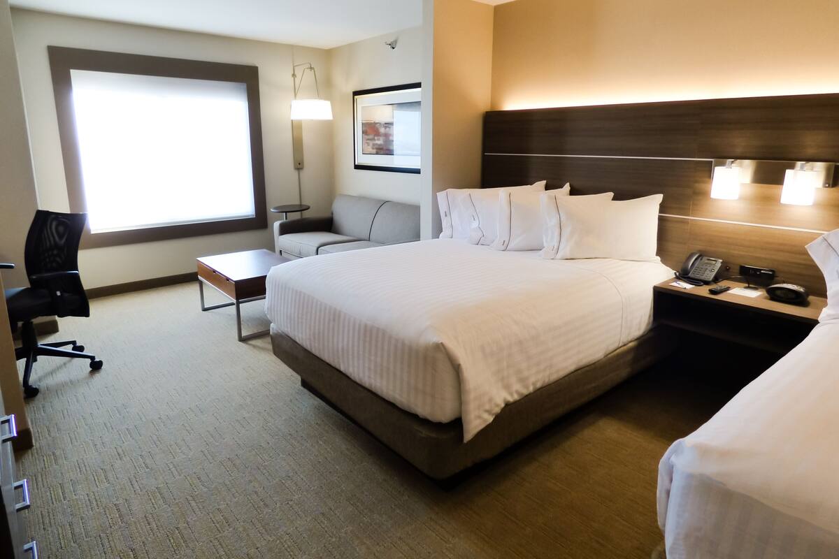 A hotel room with a large bed, a desk, a chair, a nightstand with a phone, and a window with blinds.