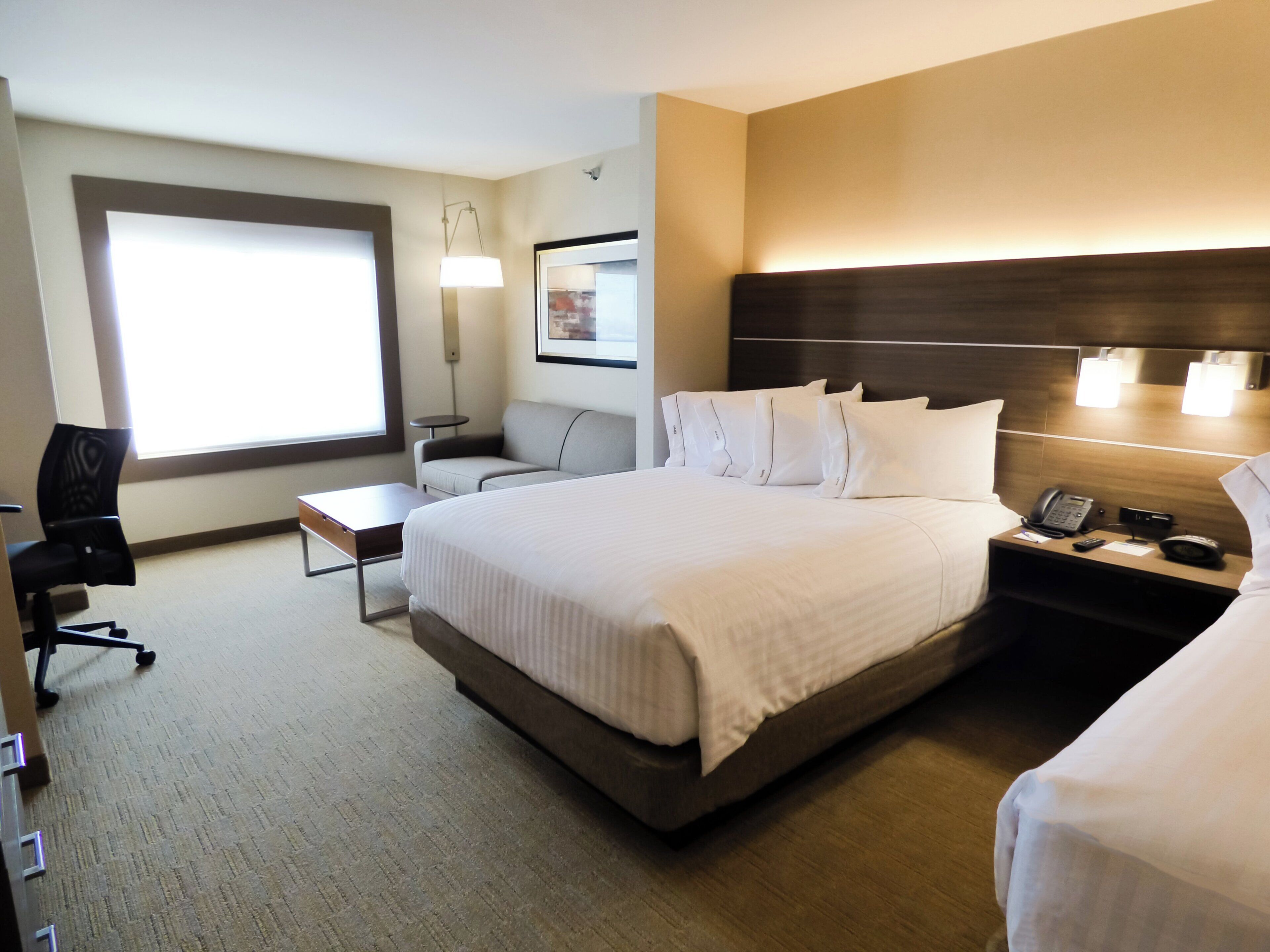 A hotel room with a large bed, a desk, a chair, and a bedside table with a phone and a lamp.