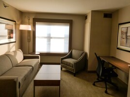Quarto de hotel com sofá, poltrona, escrivaninha, luminária e janela com persianas.