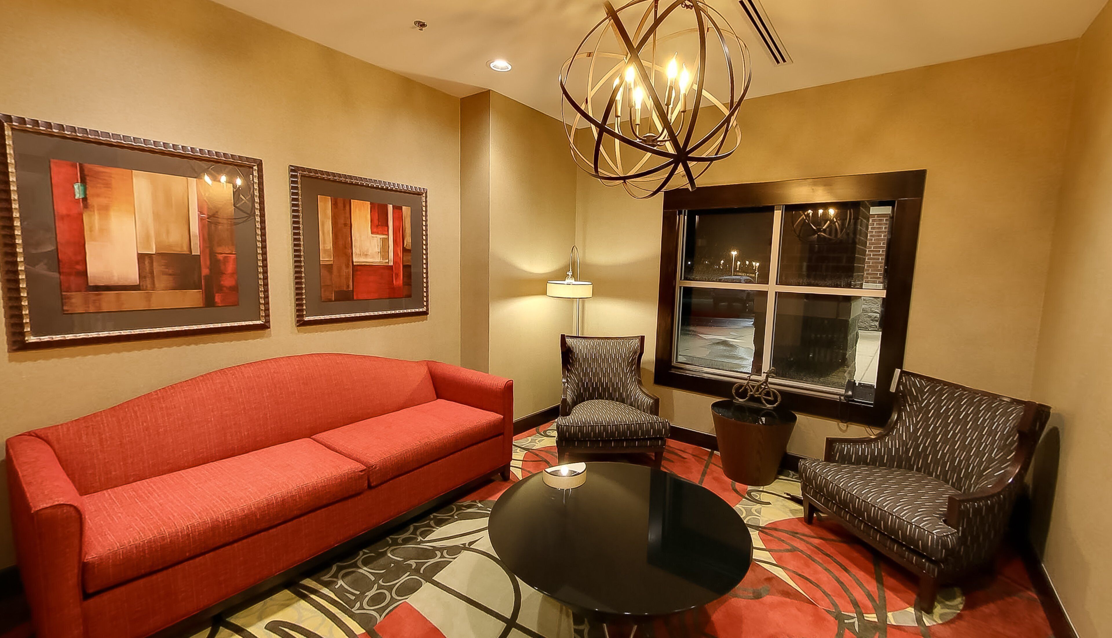 A hotel room with a red sofa, patterned armchairs, a round table, and artwork on the walls.