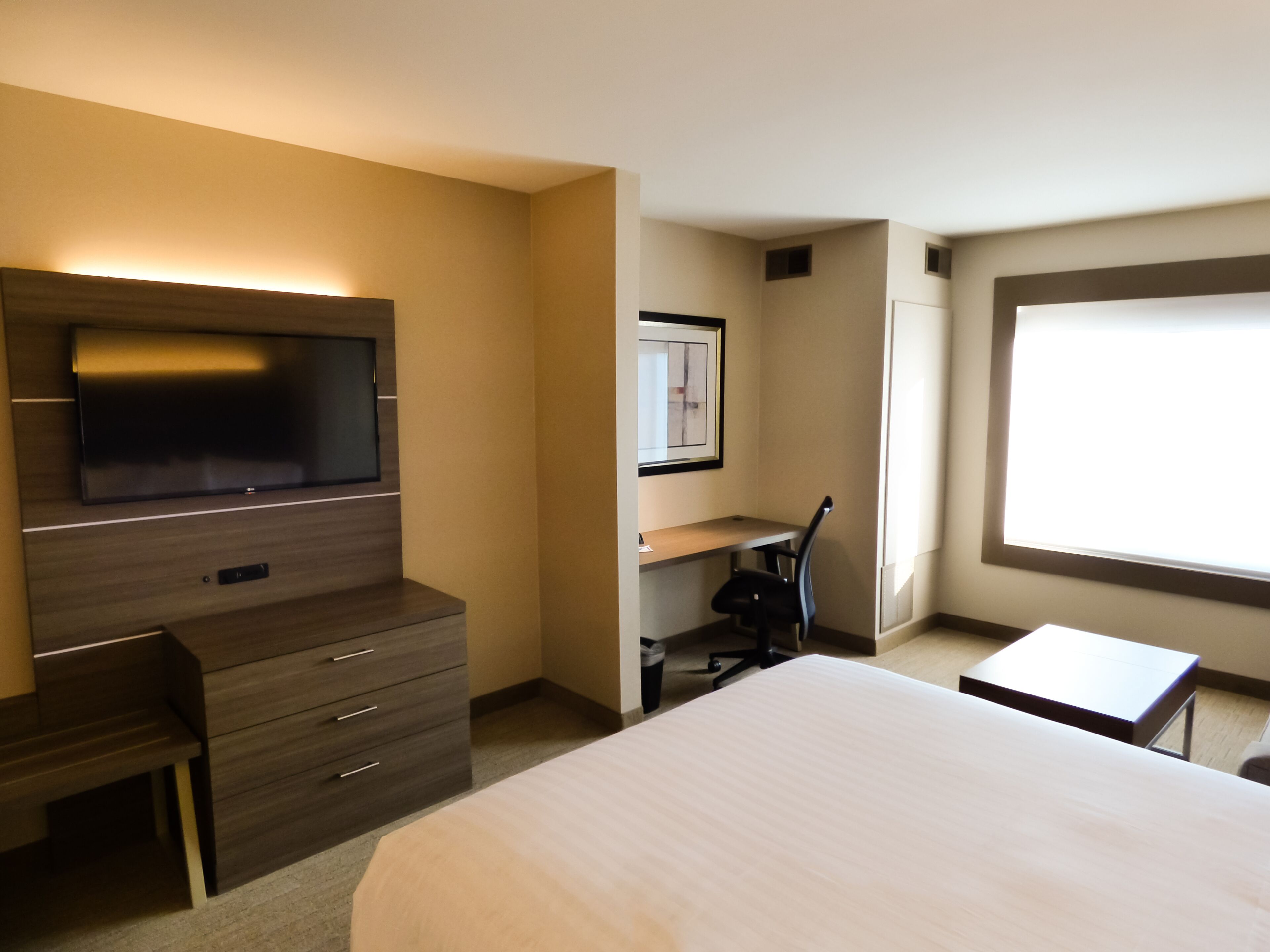 Een hotelkamer met een bed, een bureau, een stoel, een televisie en een ingelijst kunstwerk aan de muur.