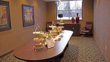 A conference room with a long table set for a buffet, two chairs, a window with a view of trees, and a vase on a cabinet.