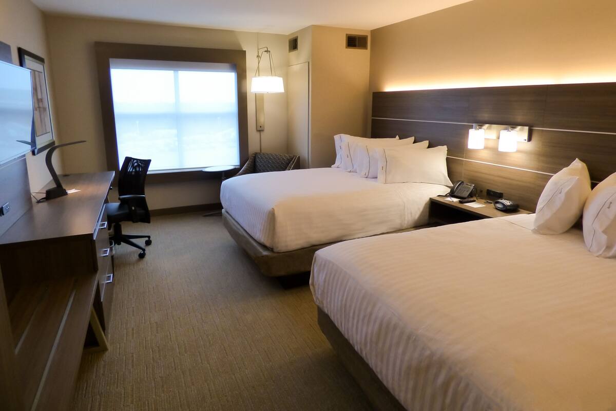 A hotel room with two beds, a desk with a computer, a chair, a window, and a nightstand with a phone and lamp.