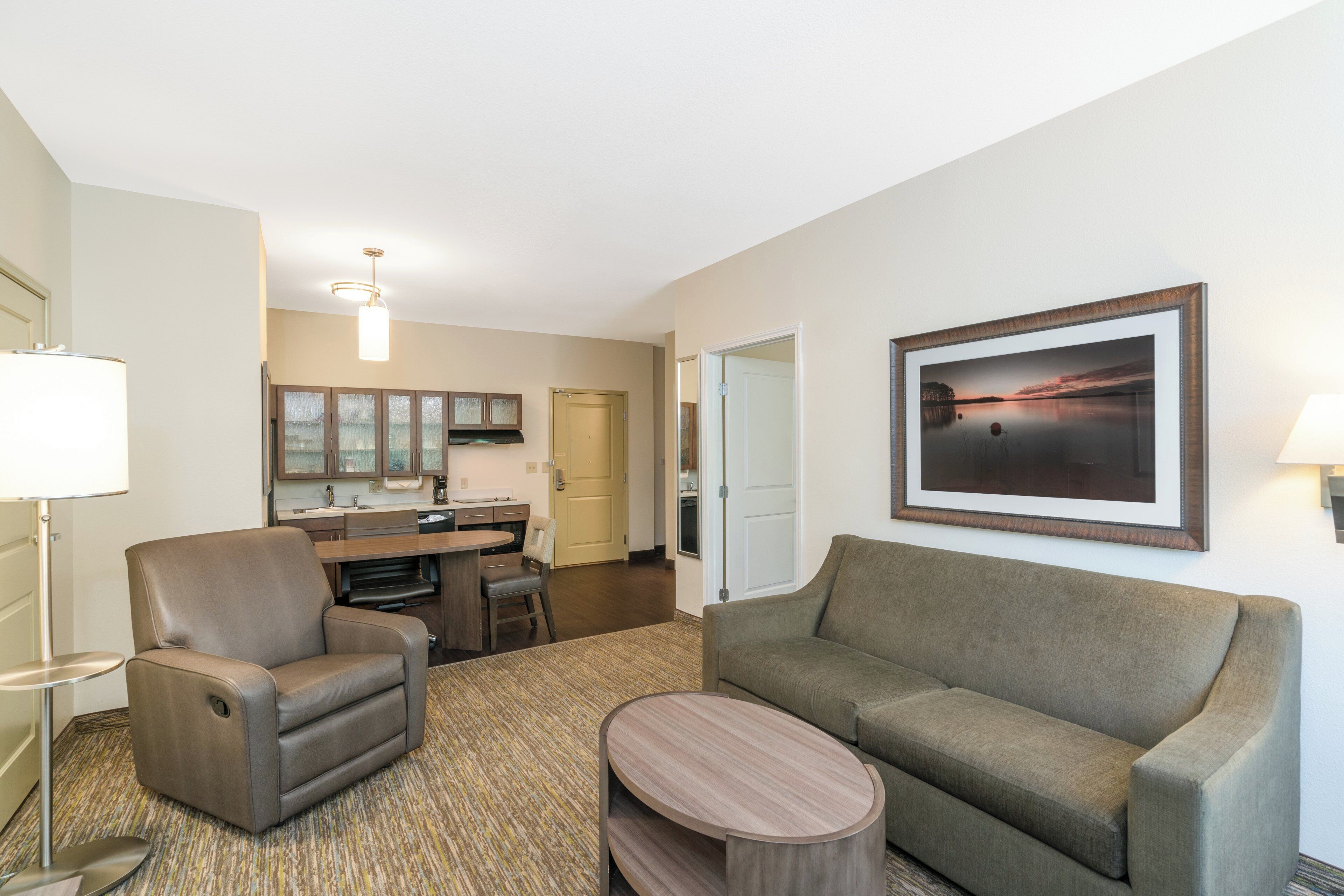 A hotel room with a sofa, armchair, dining table, and a painting on the wall.