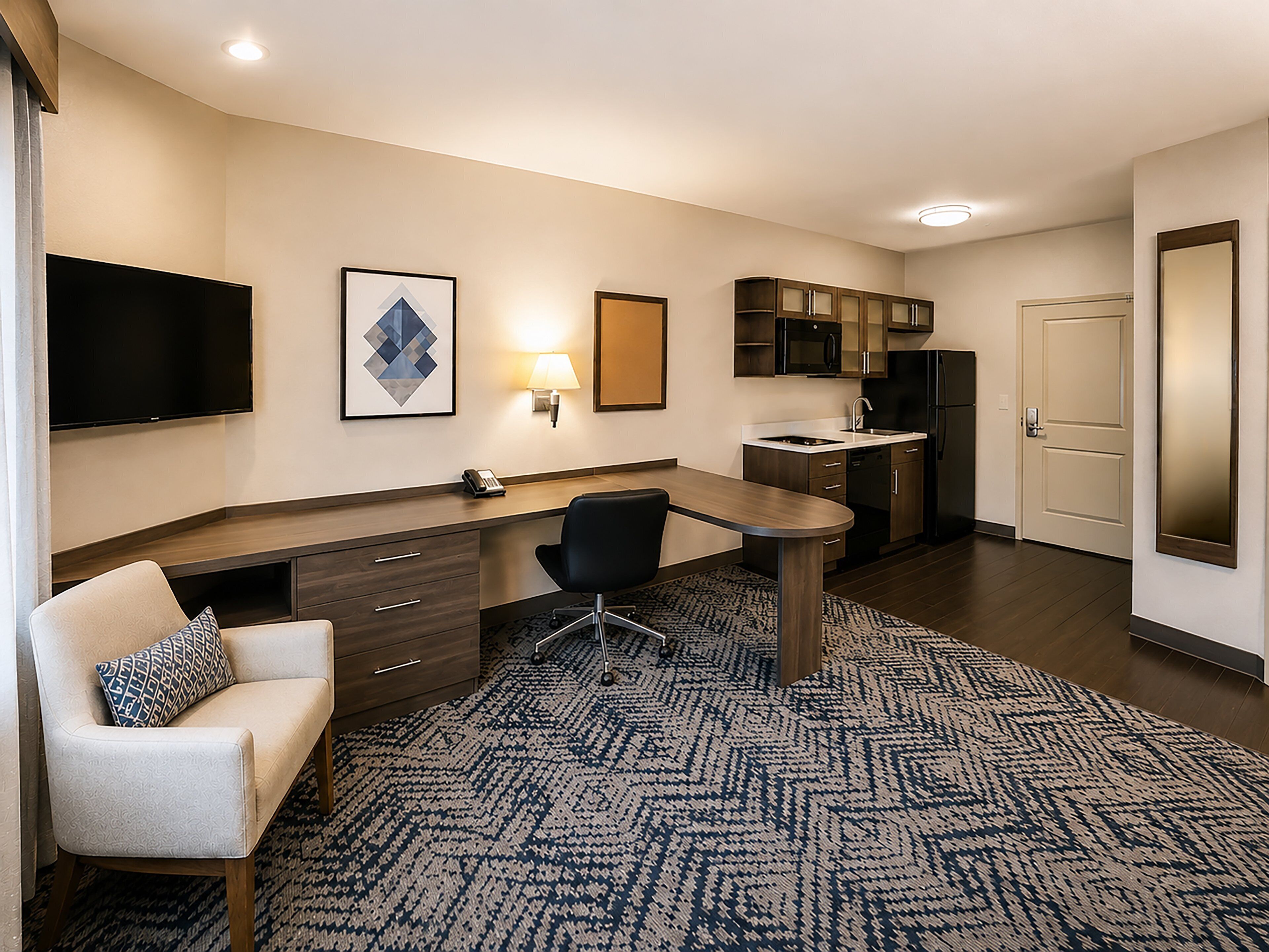 A hotel room with a flat-screen TV, a desk with a chair, a sofa, a patterned rug, a kitchen area with a microwave and refrigerator, and a door.