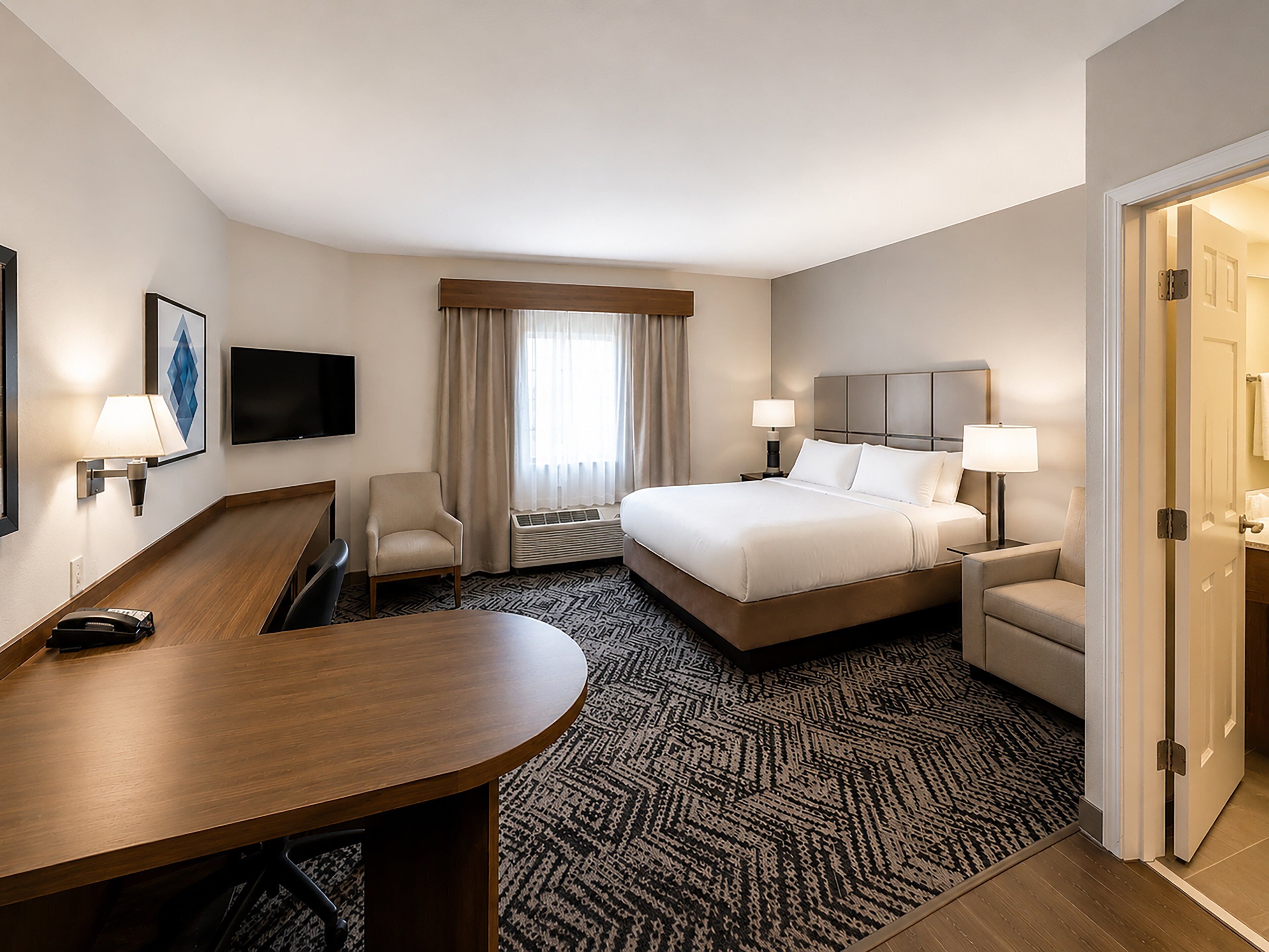 Une chambre d’hôtel comprenant un lit, deux tables de chevet avec des lampes, un bureau, une chaise, une télévision et une fenêtre avec des rideaux.