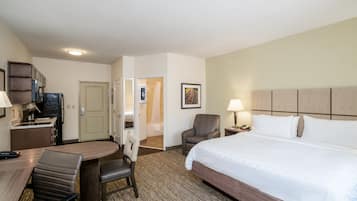 A hotel room with a bed, a desk with a chair, a small kitchenette, and a bathroom visible through an open door.