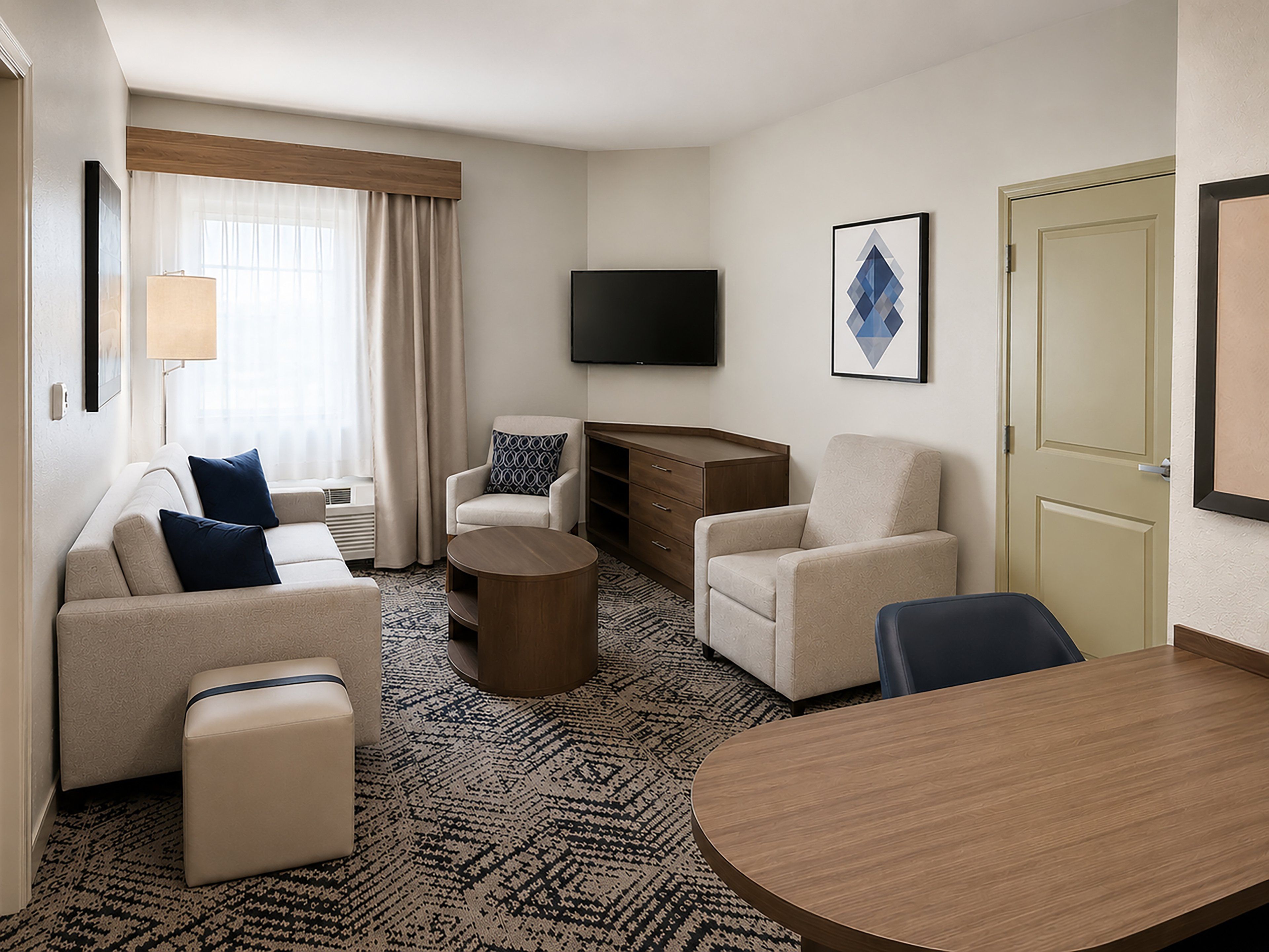 A hotel room with a sofa, armchair, ottoman, round coffee table, and a wooden side table.