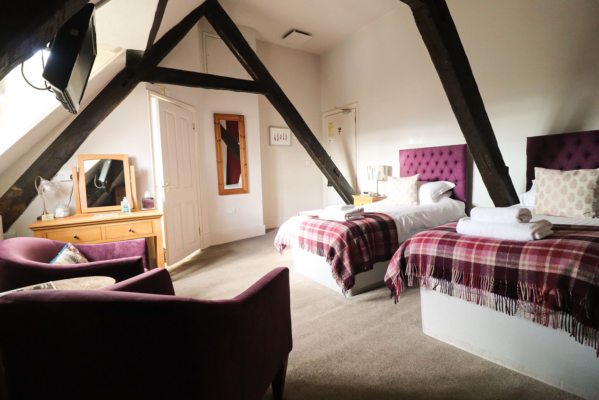 A bedroom with a sloped ceiling, a bed with a purple headboard, a desk with a mirror, and a chair.