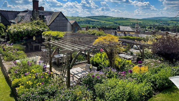 A garden with wooden pergolas and tables, surrounded by lush greenery and flowers. - The Castle Hotel (Bishops Castle)