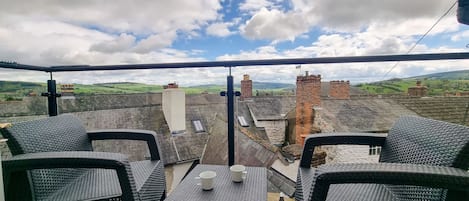A balcony with wicker furniture and a table, overlooking a village with traditional buildings and a clear sky.