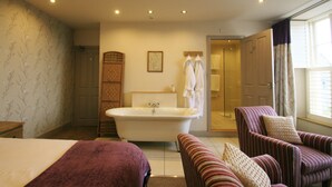 A hotel room with a large bathtub, a bed, a striped armchair, and a window with curtains. - The Castle Hotel (Bishops Castle)
