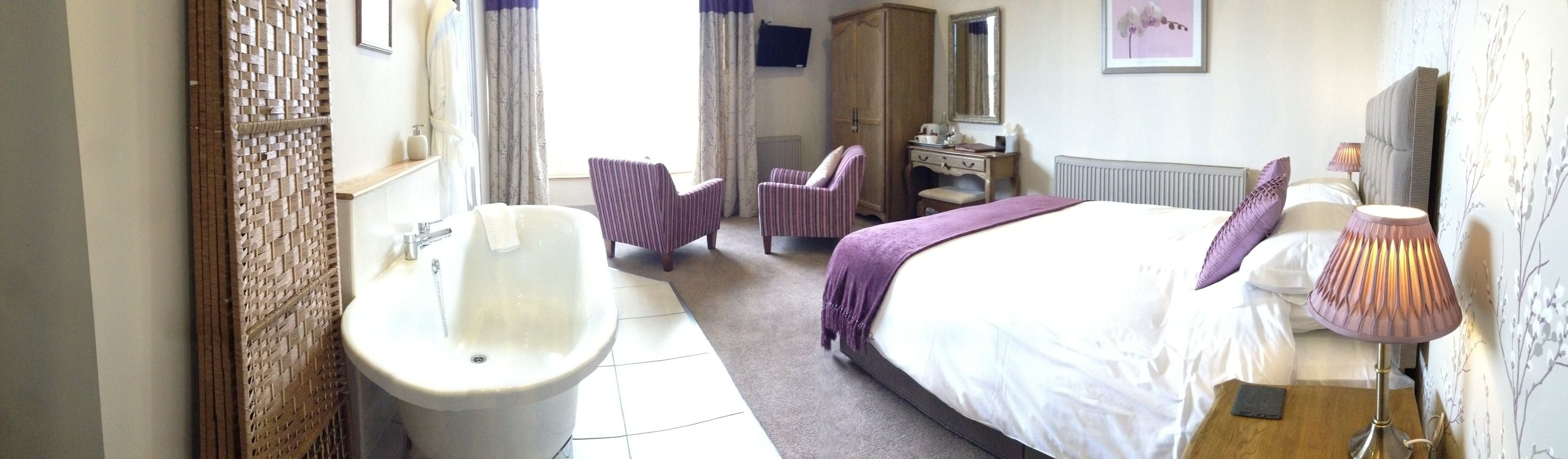 Una camera d'albergo con un letto grande, un'area salotto con una poltrona, una toeletta con uno specchio e una vasca da bagno.