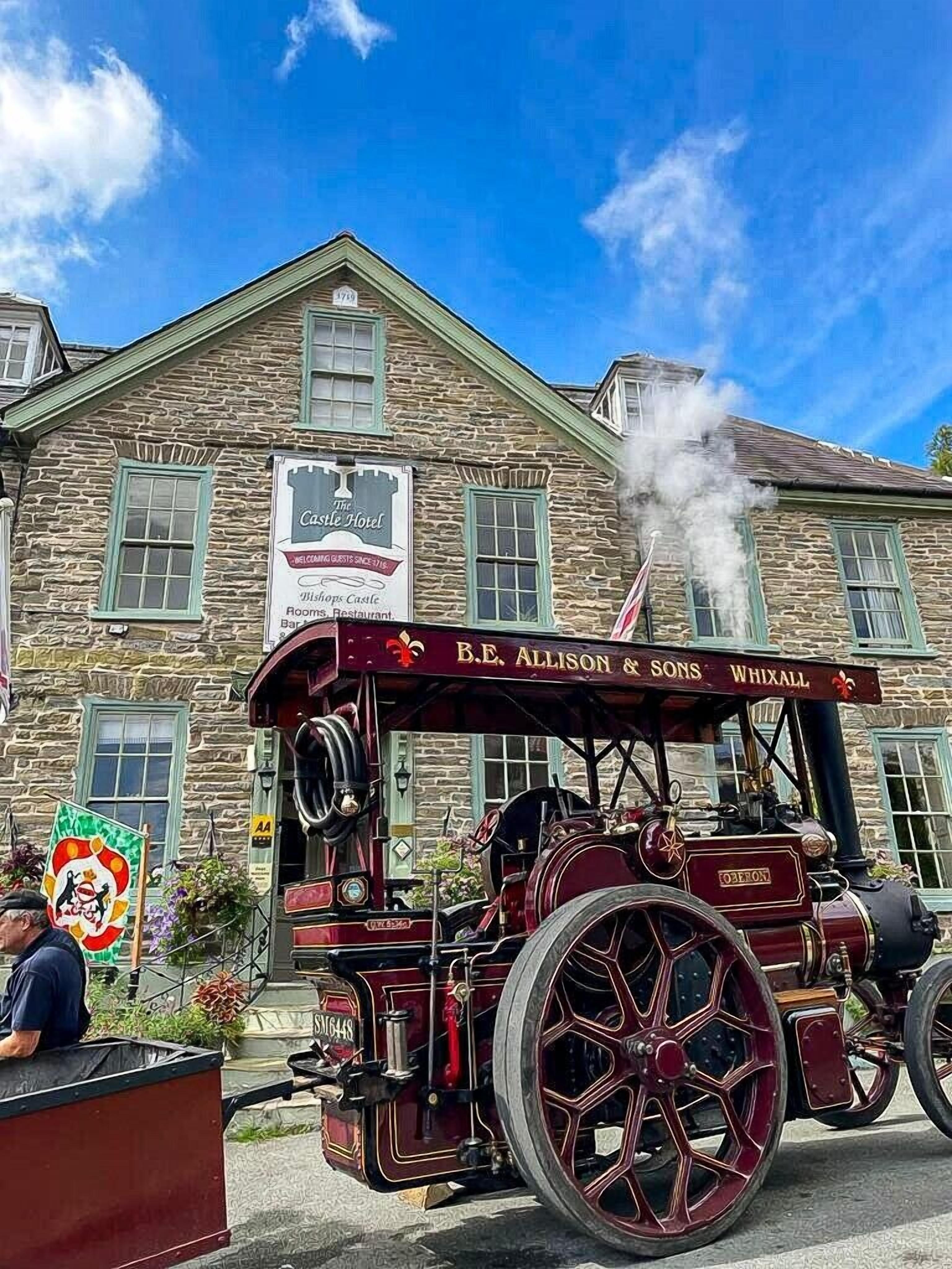 Een stoomlocomotief met de tekst "B.E. Allison & Sons Whixall" erop, geparkeerd voor een stenen gebouw met een bordje voor "Castle Hotel".