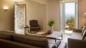 A living room with a sofa, armchair, coffee table, and a view of the sea through a glass door. - Aeolis Tinos Suites (Tinos)