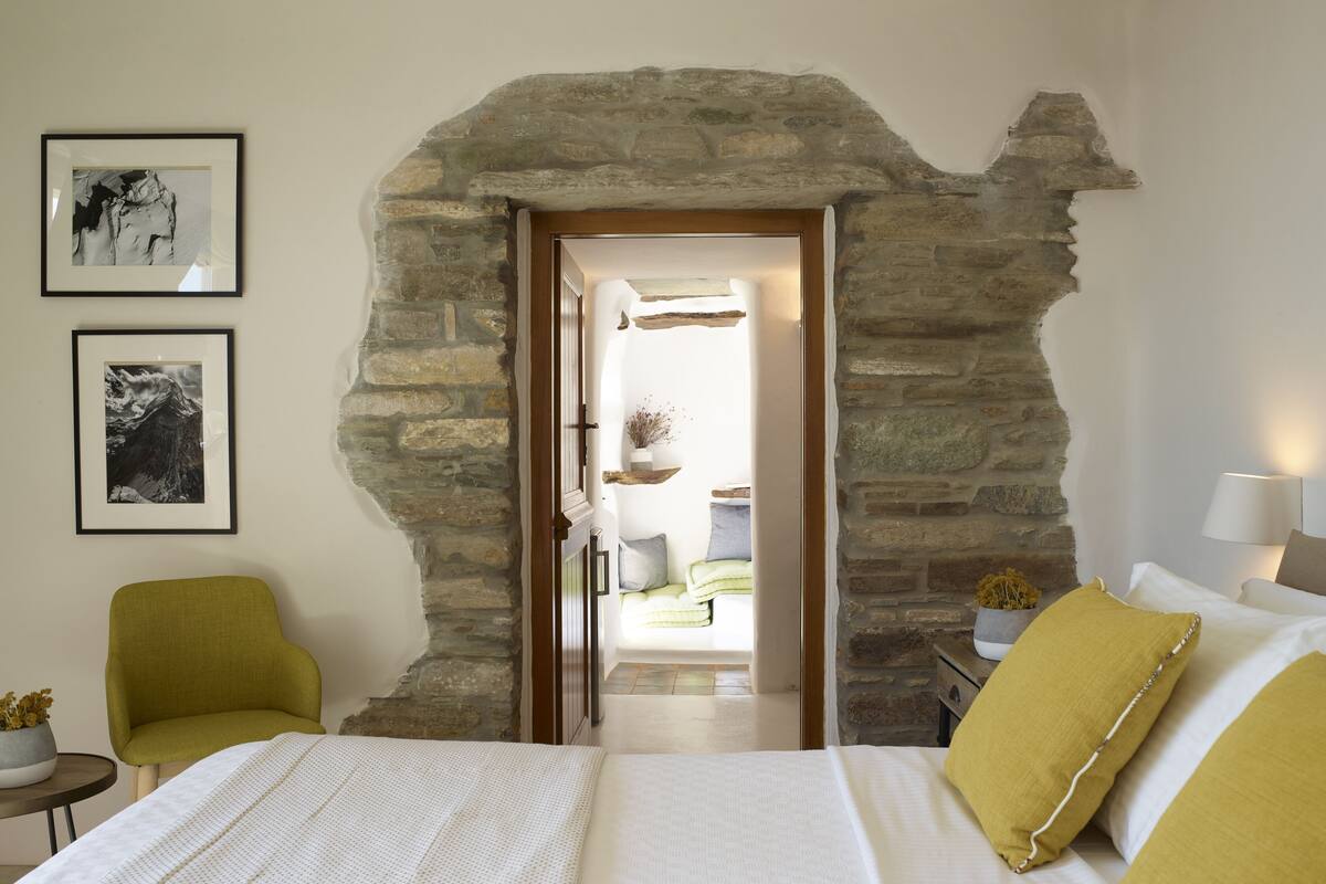 A bedroom with a stone archway, a bed with white bedding, a green armchair, and two framed pictures on the wall.