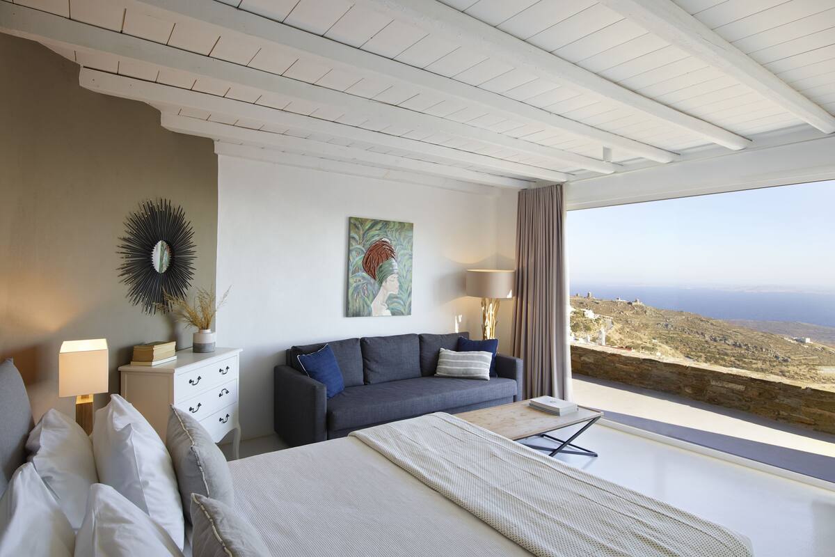 A modern bedroom with a large bed, a sofa, a desk, and a view of the landscape.