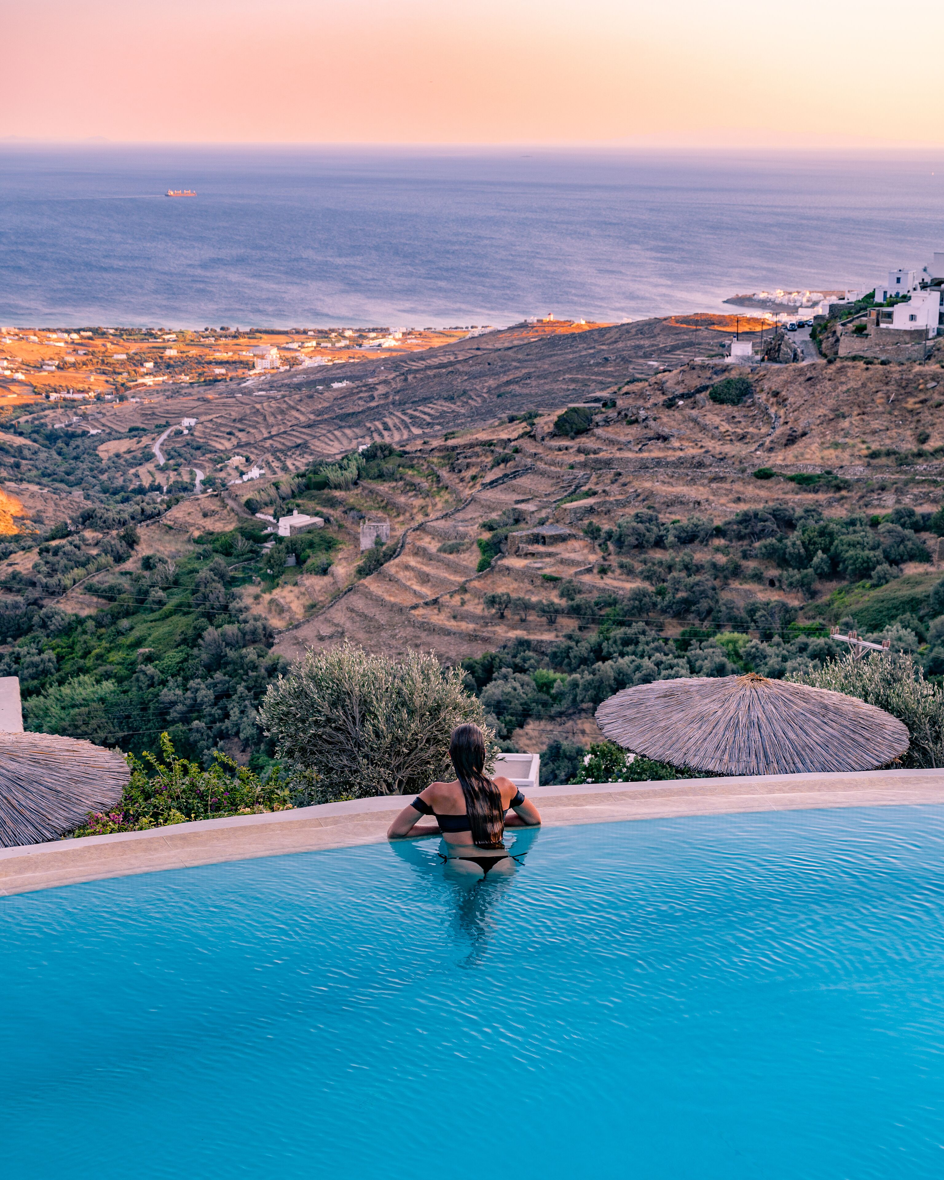A person relaxing in a pool with a view of the sea and a hilly landscape.