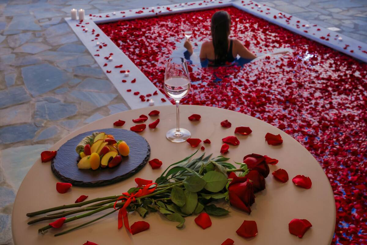 A terrace with a table set for two, surrounded by rose petals, a glass of wine, and a fruit arrangement.