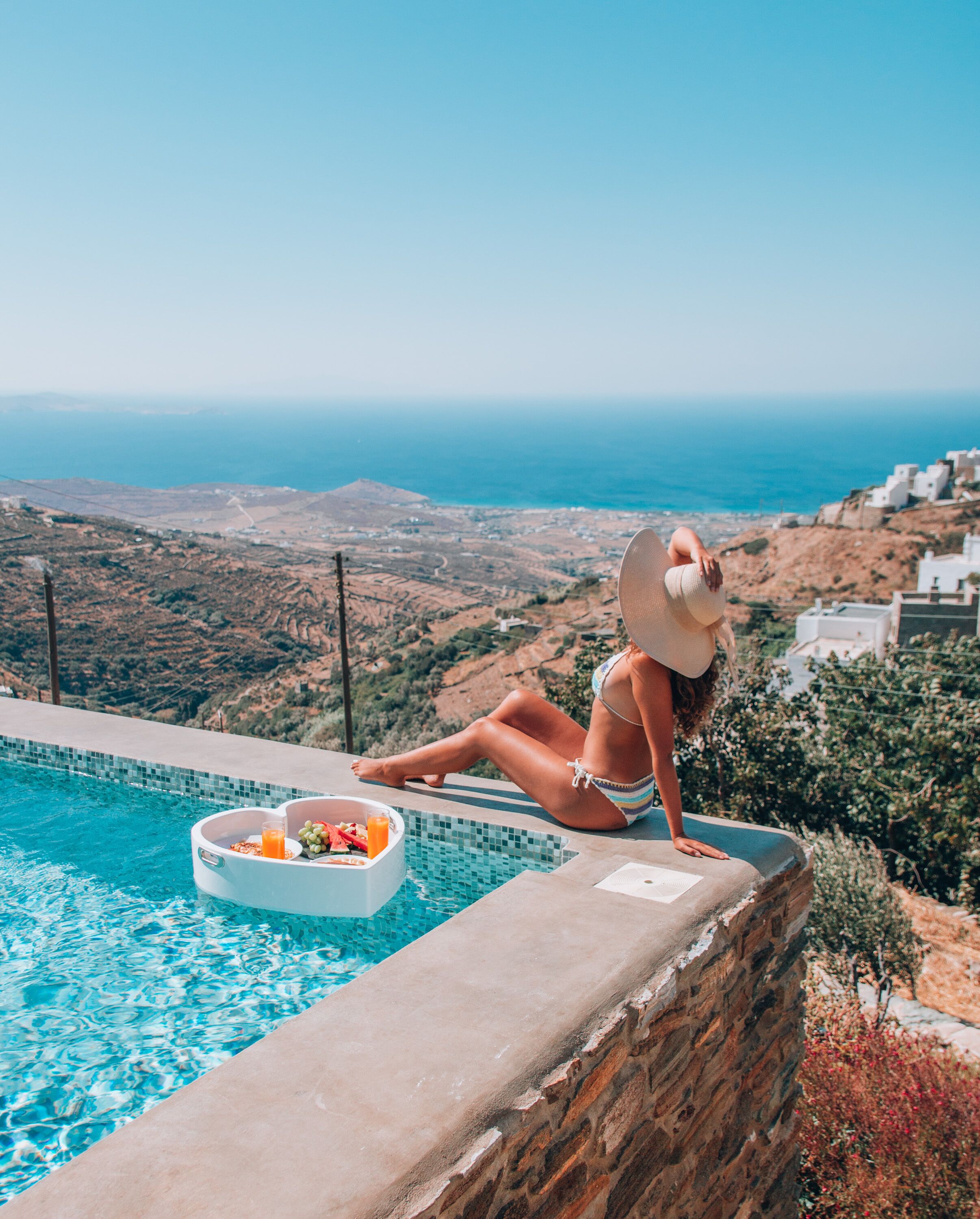 A person sitting by a pool with a view of the sea and hills.
