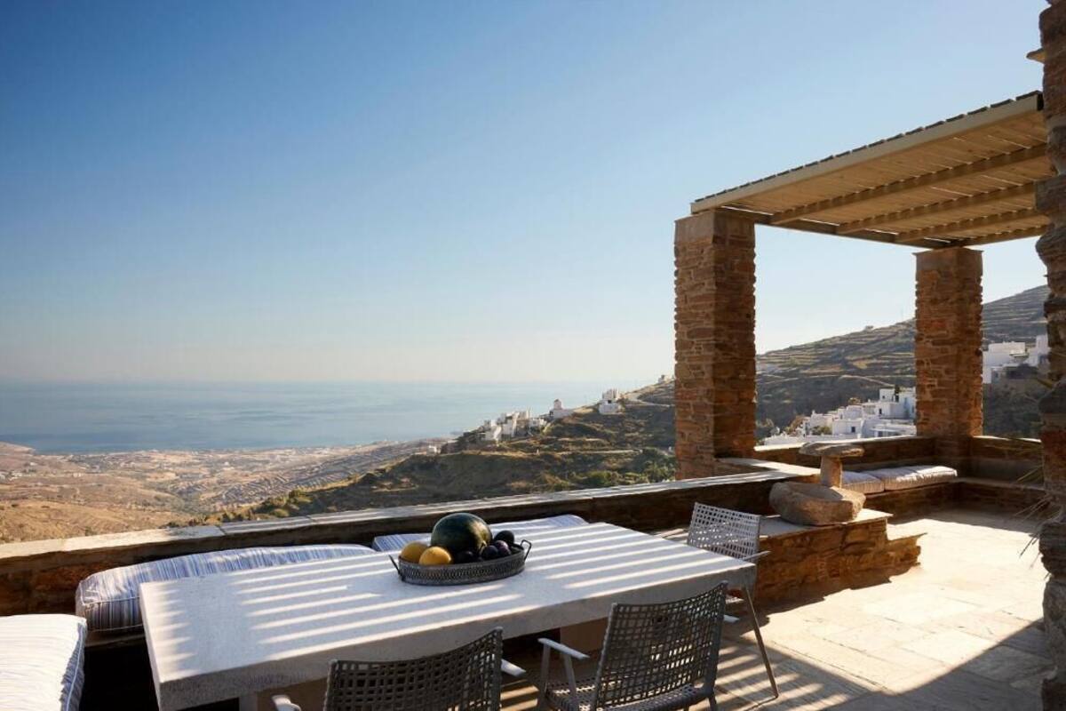 An outdoor terrace with a table and chairs, a stone pergola, and a view of the coastline.