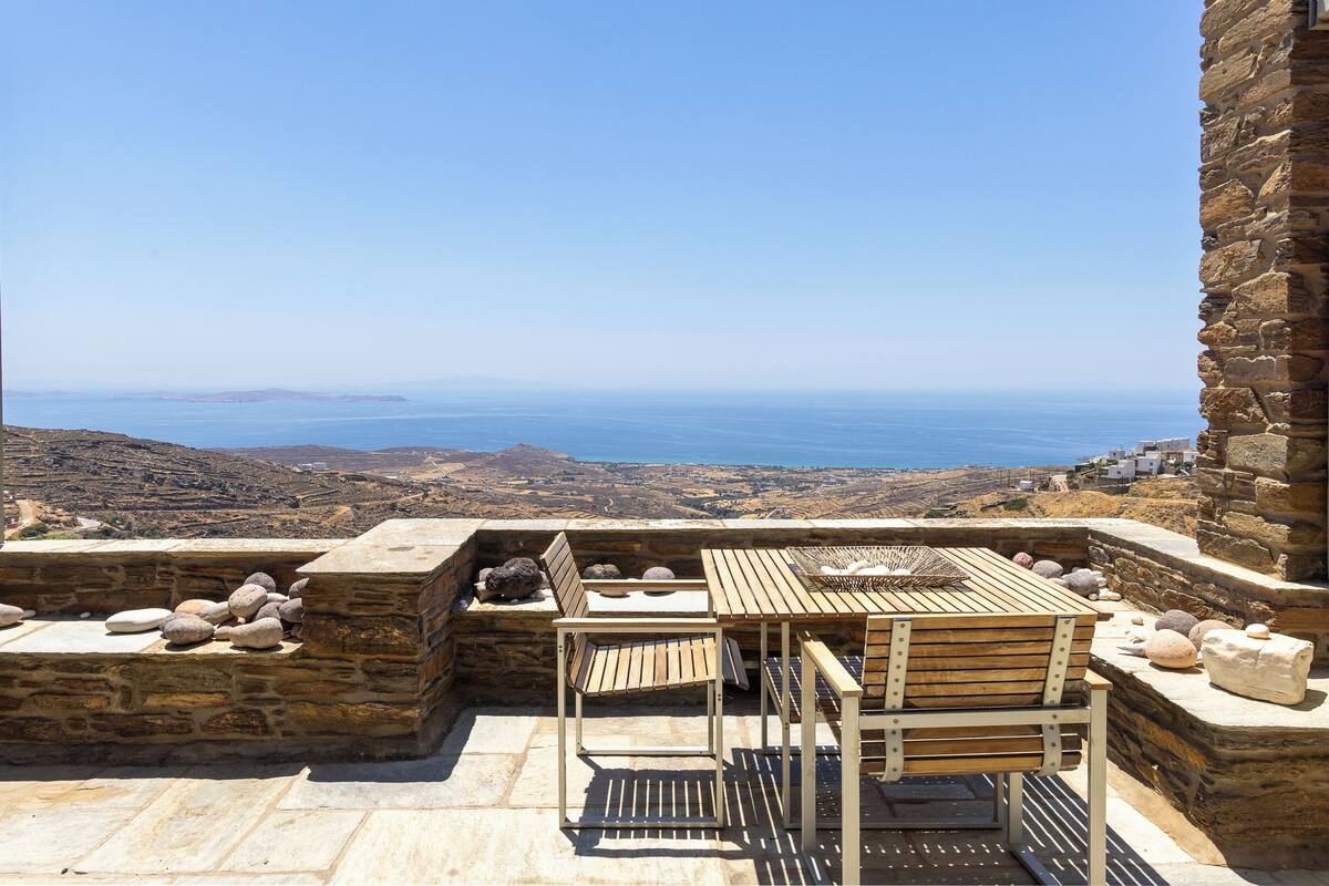 A stone patio with a wooden table and chairs overlooking a coastal landscape.