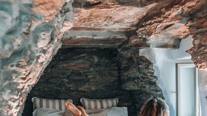 A bedroom with a stone wall, a bed with striped pillows, and a window. - Aeolis Tinos Suites (Tinos)