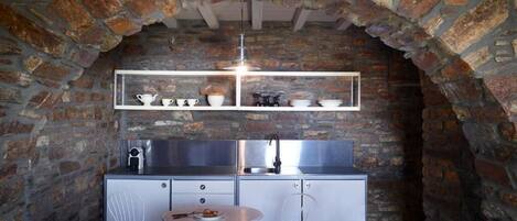 A rustic kitchen with exposed brick walls, a metal sink, and a round table with a bowl of fruit.