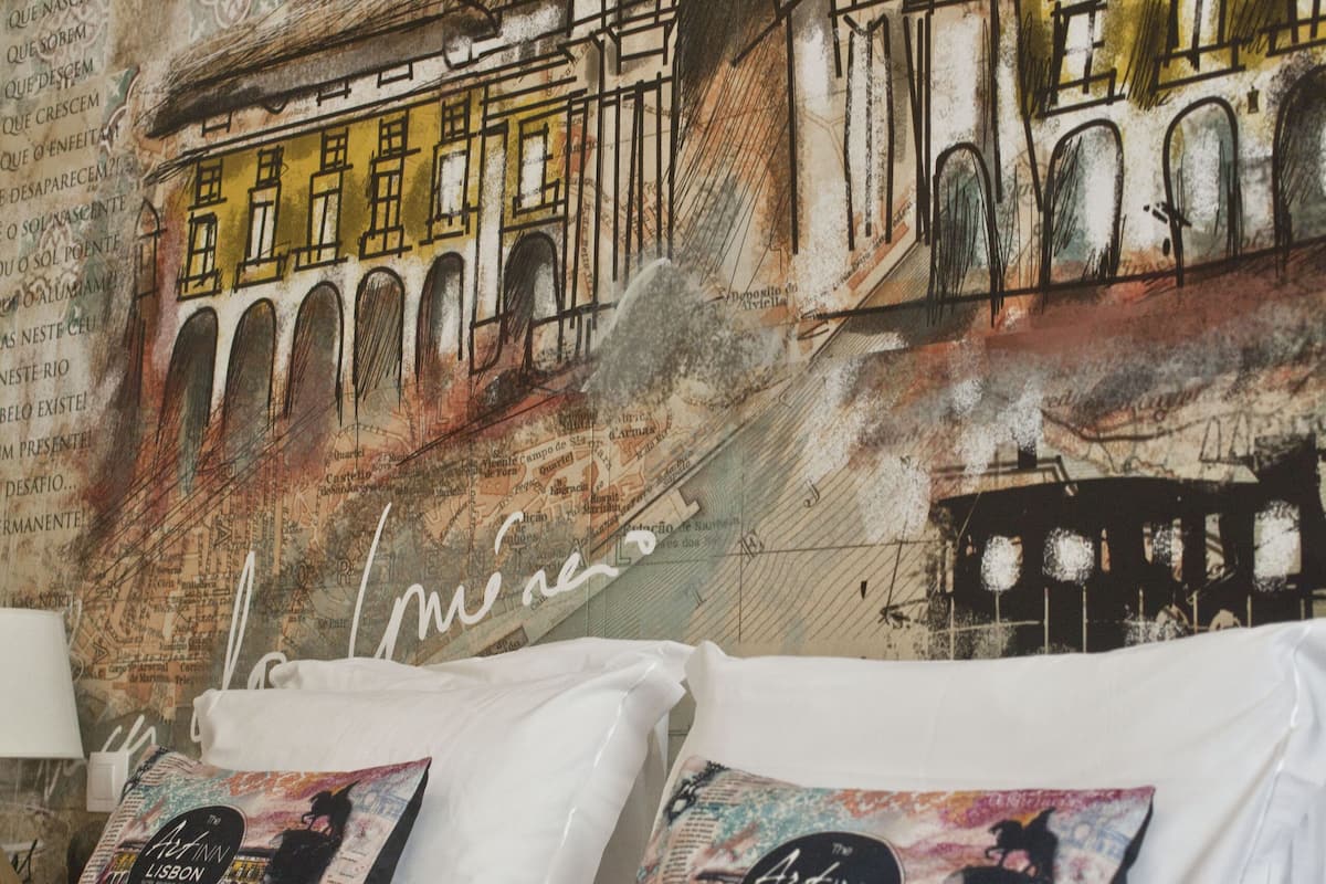 Uma cama com lençóis brancos e duas almofadas, uma com desenho de paisagem urbana e outra com capa de livro.
