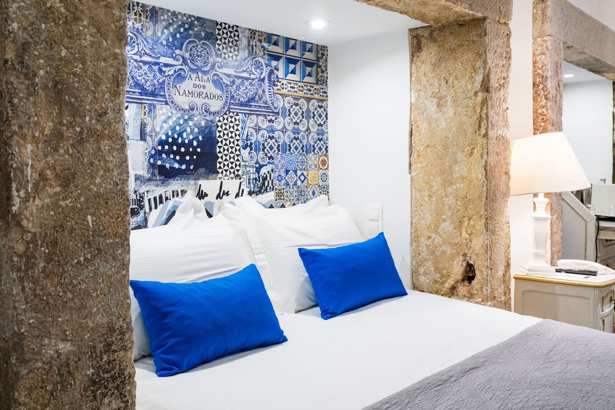 Cama com roupa de cama branca e duas almofadas azuis, parede com design estampado em azul e branco, e um pilar de pedra.