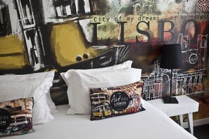 Una cama con almohadas blancas y un cojín decorativo de colores, una mesita de noche blanca con una lámpara, y un mural en la pared que representa la arquitectura de Lisboa.